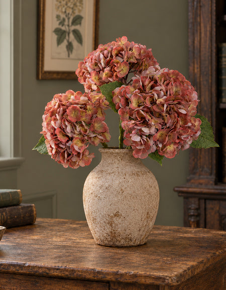Three antique brick rose hydrangeas in a stoneware vase beside books in a vintage study.