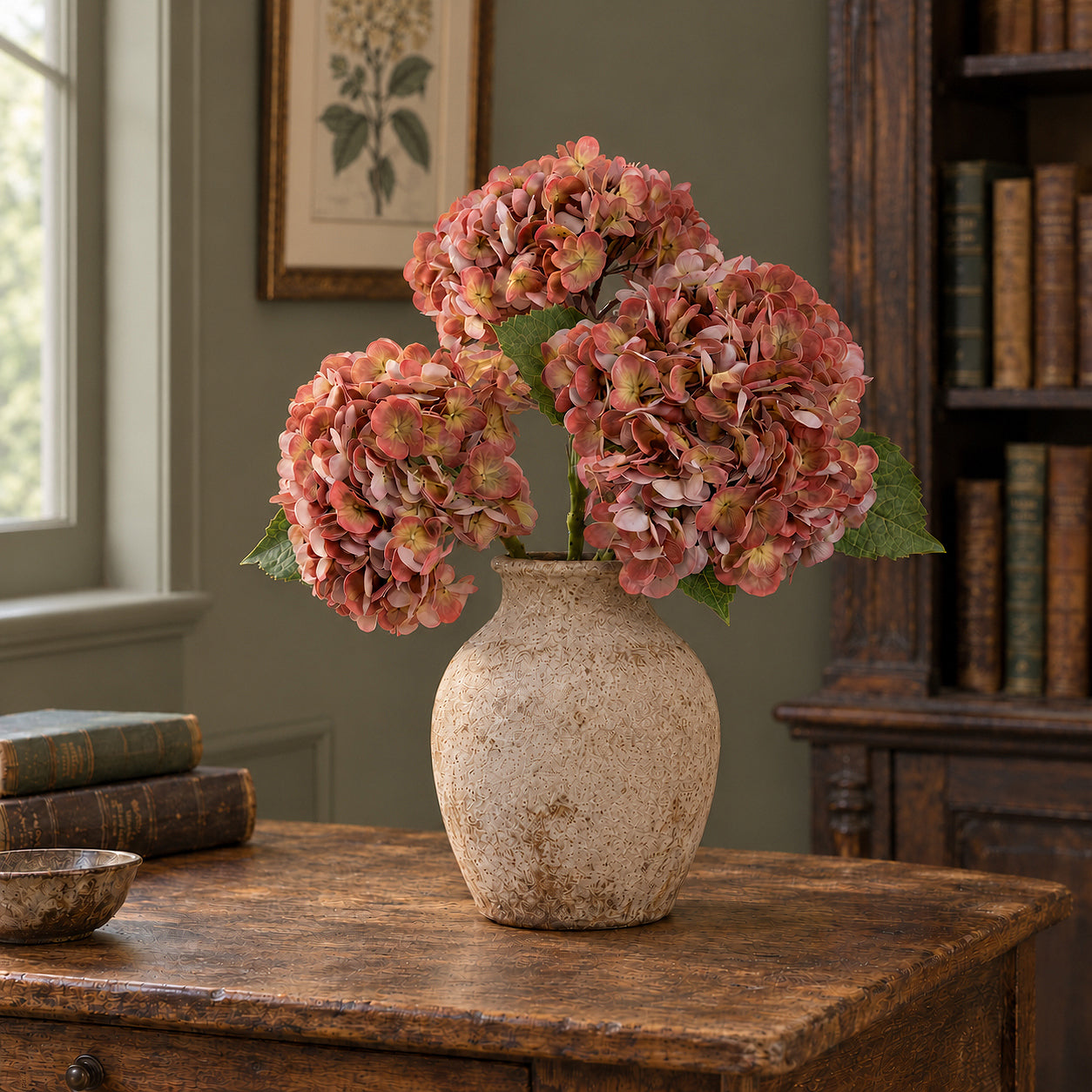 Three antique brick rose hydrangeas in a stoneware vase beside books in a vintage study.