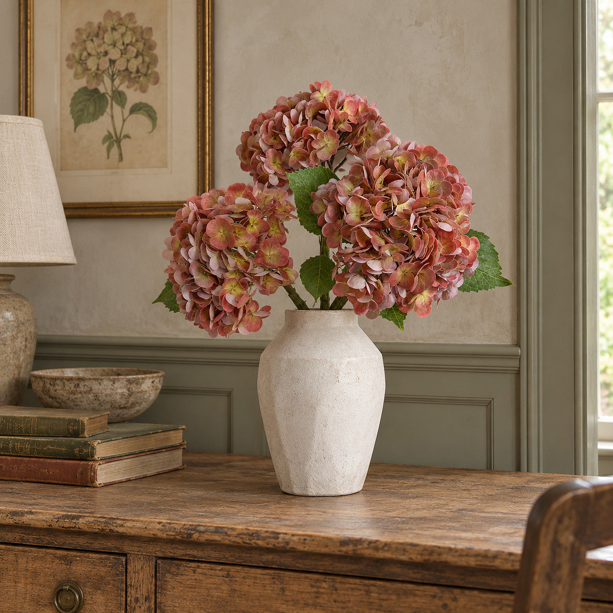 Three antique brick rose hydrangeas in a pale vase on a wood sideboard beneath botanical artwork.