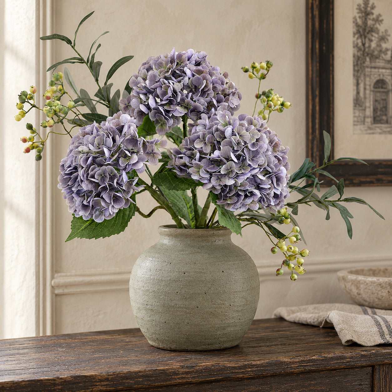 Three antique lavender hydrangeas with leafy branches and berries in a gray-green vase on a rustic table.