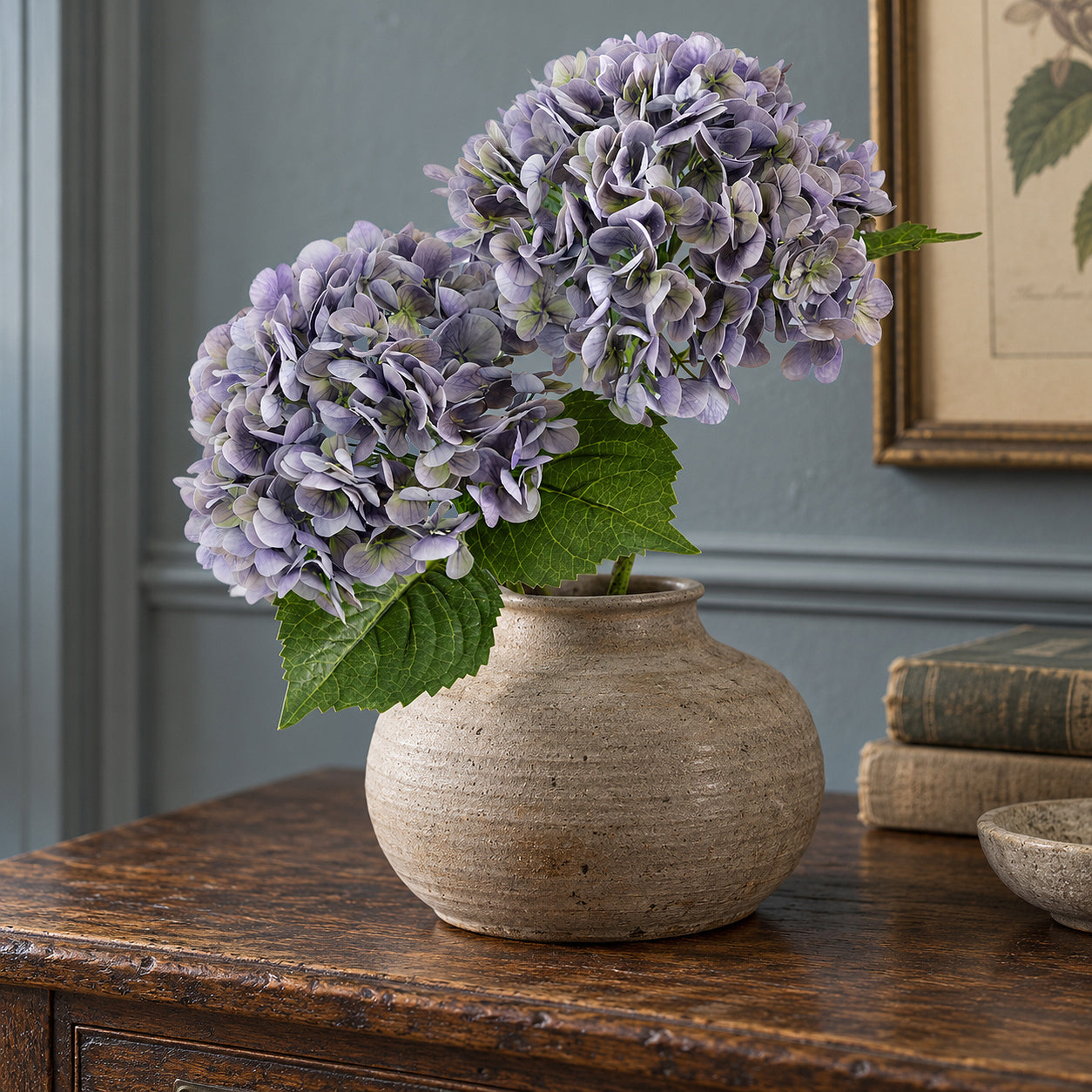 Two antique lavender hydrangeas in a rounded stoneware vase on a wood table.