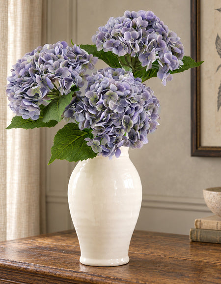Three antique lavender hydrangeas in a cream vase on an oak sideboard.