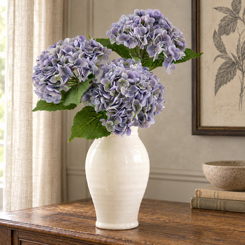 Three antique lavender hydrangeas in a cream vase on an oak sideboard.