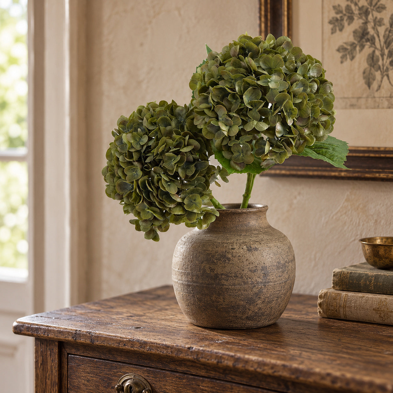 Two antique olive hydrangeas in a rounded brown stoneware vase on a wood table by a window.