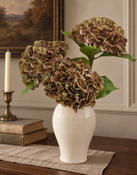 Three antique russet hydrangeas in a cream vase on a walnut console beside books and a candle.
