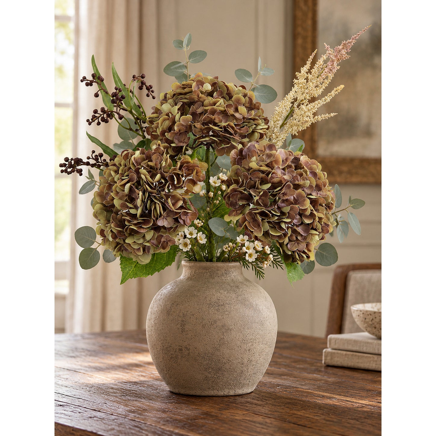 Three antique russet hydrangeas with berry stems and greenery in a stoneware vase on a dining table.