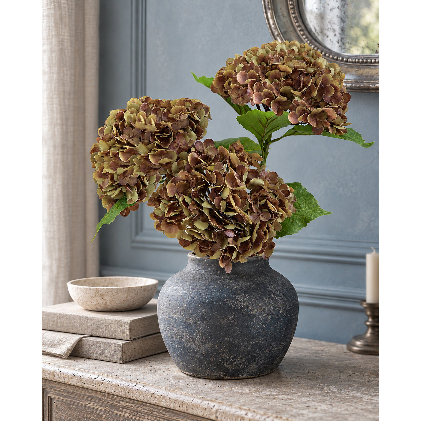Three antique russet hydrangeas in a dark rounded vase on a console against a blue-gray wall.