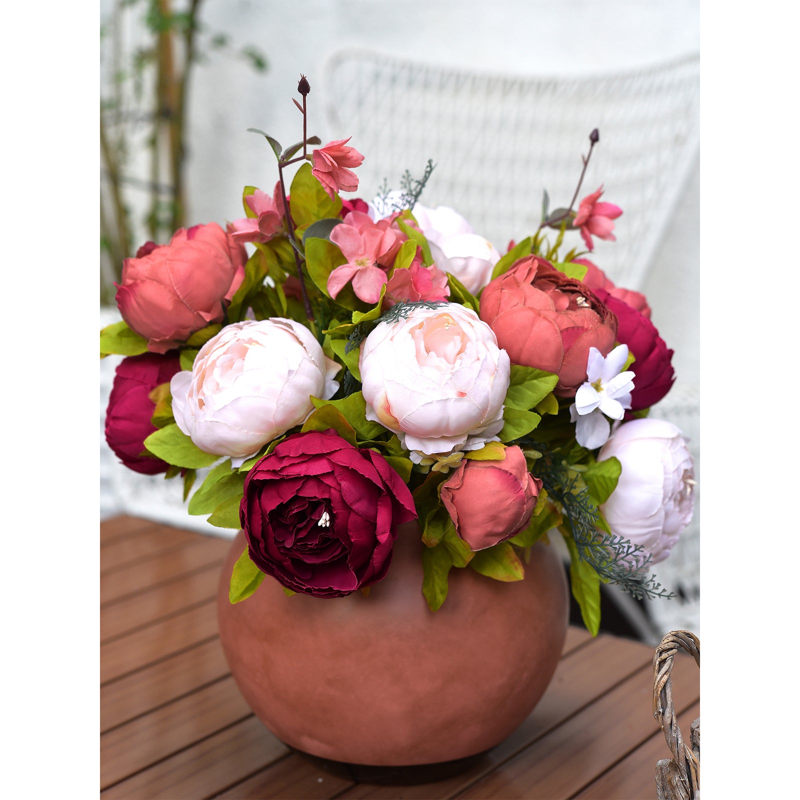 Artificial peony arrangement placed in a rustic terracotta vase on a wooden table, featuring soft pink, coral, and deep red tones.