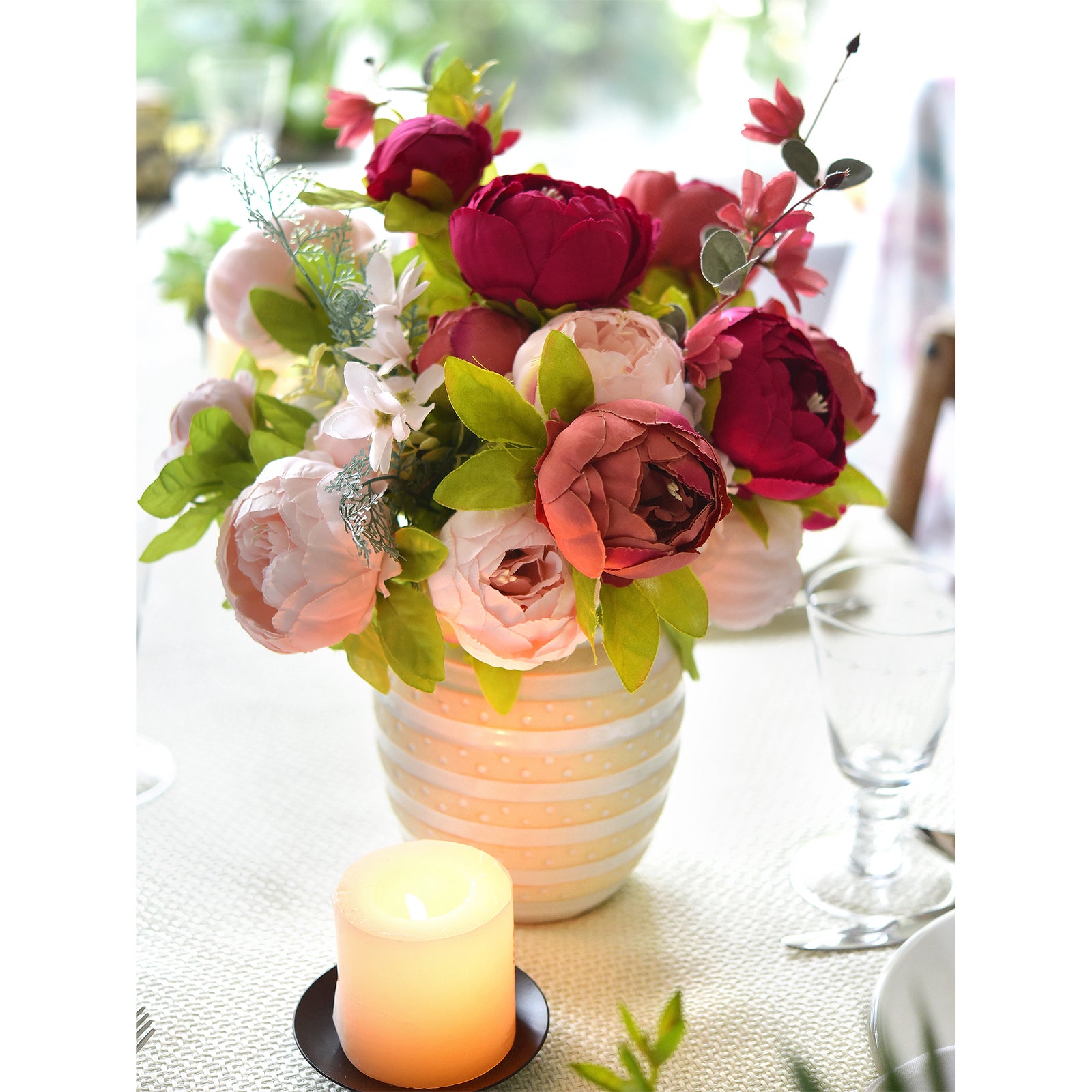 Artificial peony bouquet arranged in a cream-colored vase, styled with a lit candle and glassware for an elegant table setting.