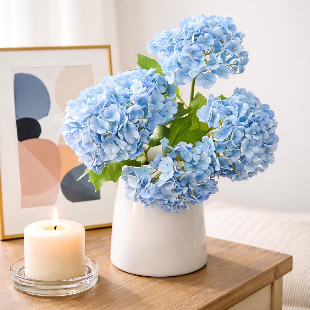 Blue real touch hydrangea stems arranged in a white vase on a wooden table beside a candle.