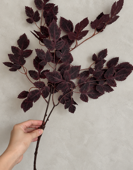 Hand holding a burgundy artificial maple branch against a light gray wall.