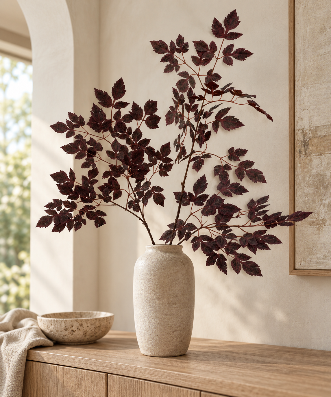 Burgundy foliage arranged in an ivory vase beside a bowl on a wooden cabinet.