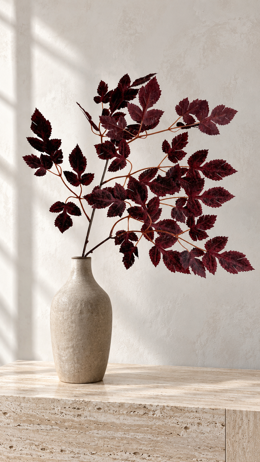 Burgundy maple foliage in a narrow beige vase on a sunlit stone surface.