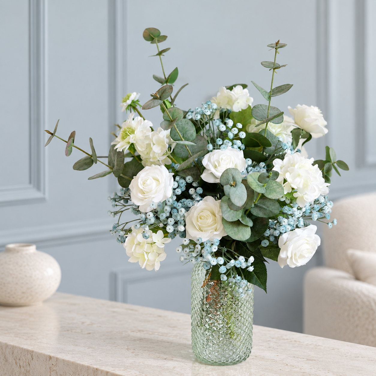 White flowers and dusty blue baby's breath with eucalyptus in a textured glass vase against blue wall paneling.