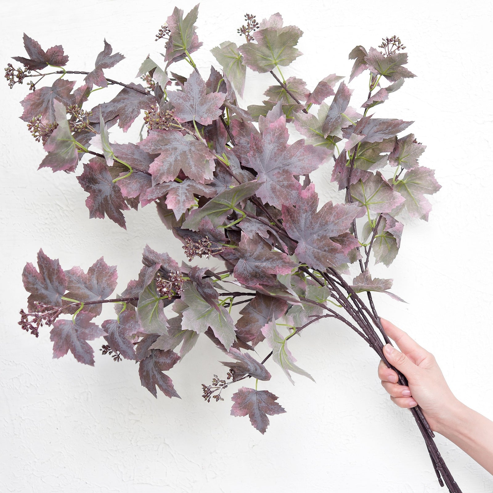 Hand holding a group of dusty mauve brown artificial maple leaf stems against a white wall.