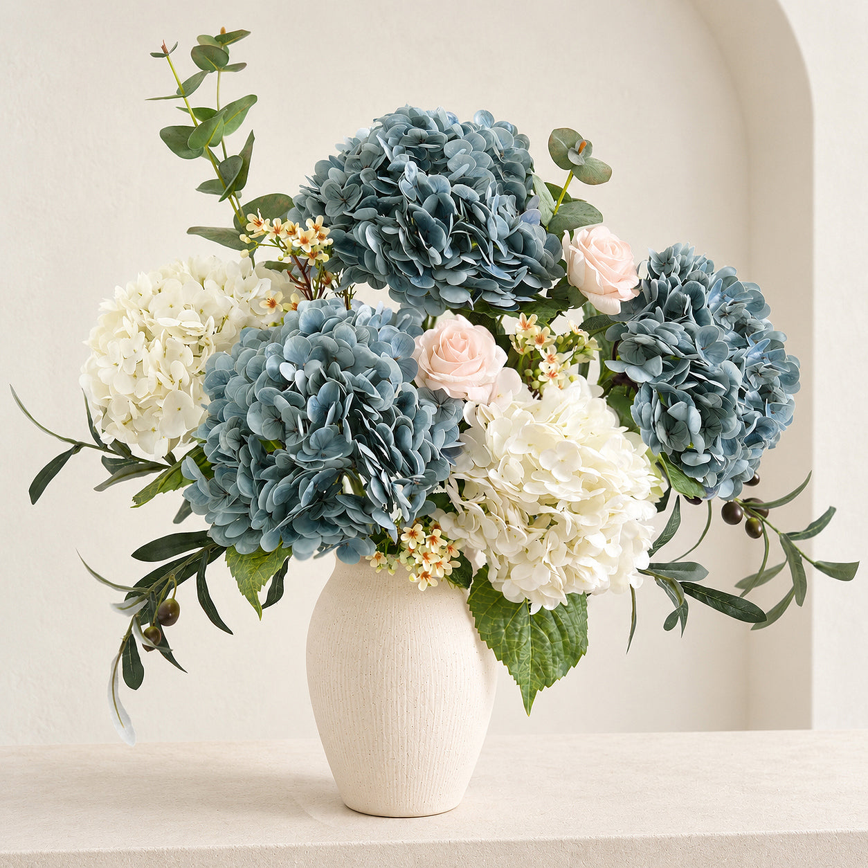 Three dusty slate blue hydrangeas with white flowers, blush roses and greenery in a cream vase.