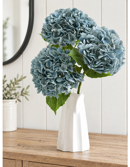 Three dusty slate blue hydrangea stems arranged in a white sculptural vase on a wood cabinet.