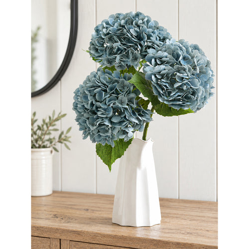 Three dusty slate blue hydrangea stems arranged in a white sculptural vase on a wood cabinet.
