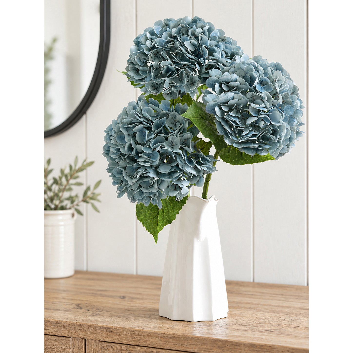 Three dusty slate blue hydrangea stems arranged in a white sculptural vase on a wood cabinet.