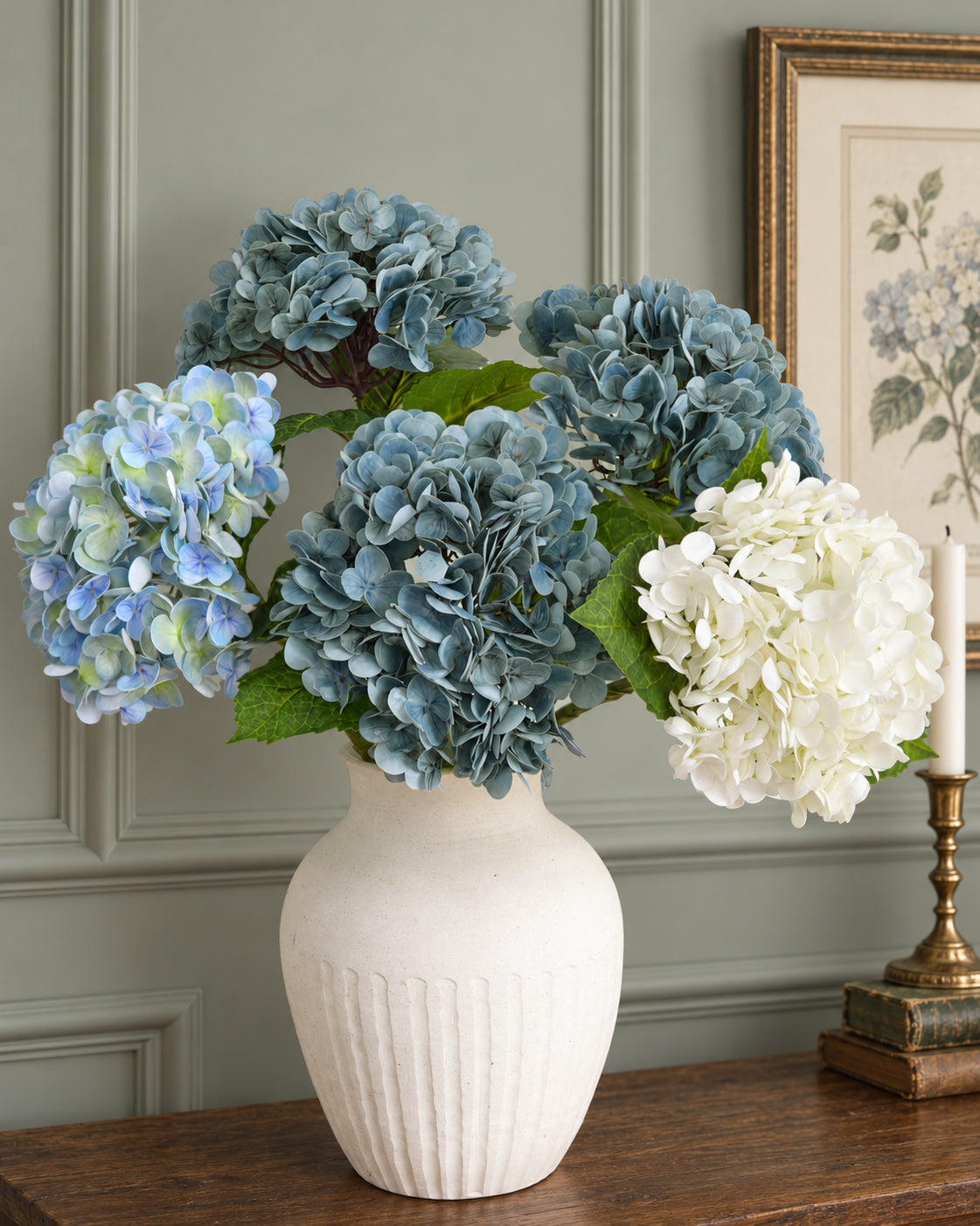 Dusty slate blue hydrangeas with pale blue and white hydrangeas in a white vase against a green wall.