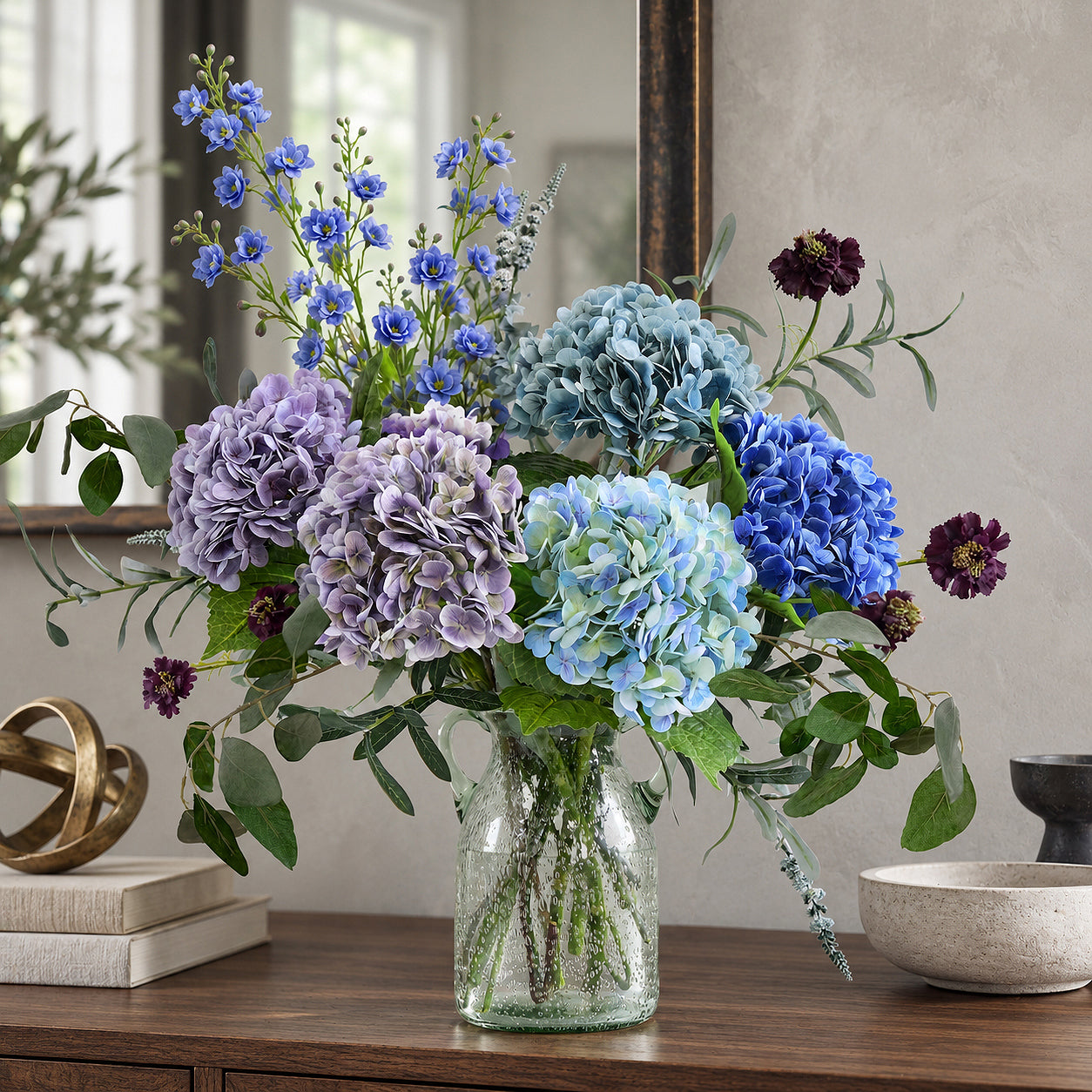 Dusty slate blue hydrangeas with lavender and blue flowers in a clear glass vase on a walnut console.