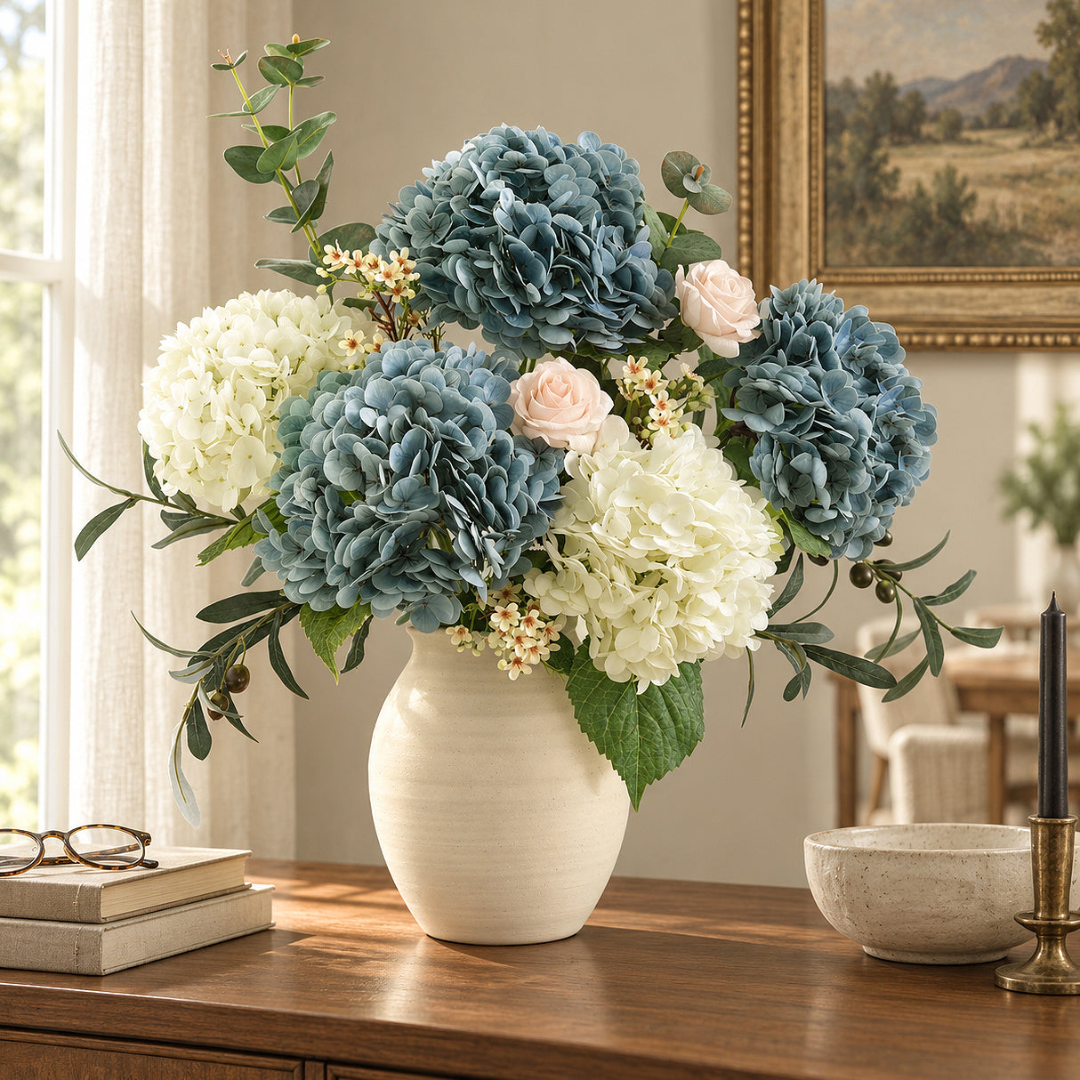 Three dusty slate blue hydrangeas with white flowers and blush roses in a cream vase on a wooden sideboard.