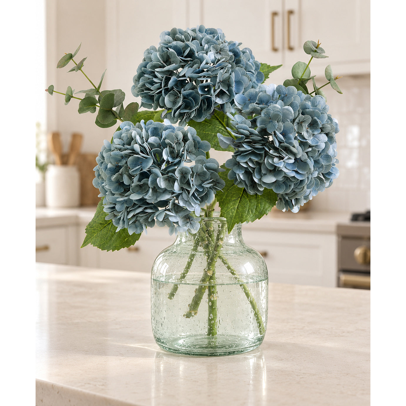 Three dusty slate blue hydrangeas with greenery in a clear glass vase on a kitchen counter.
