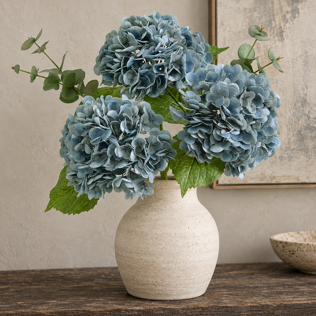 Three dusty slate blue hydrangeas with greenery in a rounded stoneware vase.