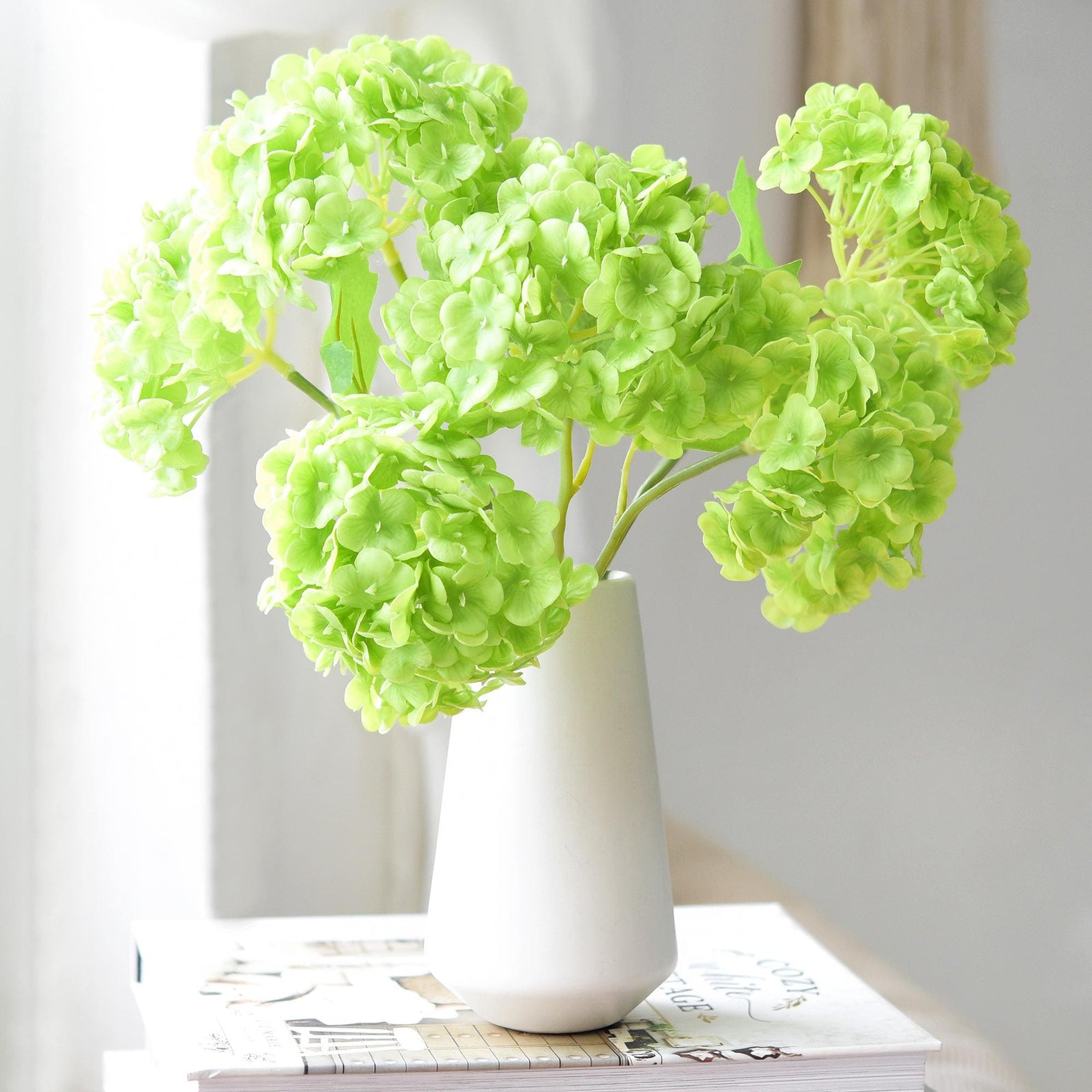 Lime green real touch hydrangea stems arranged in a matte white vase on stacked books.