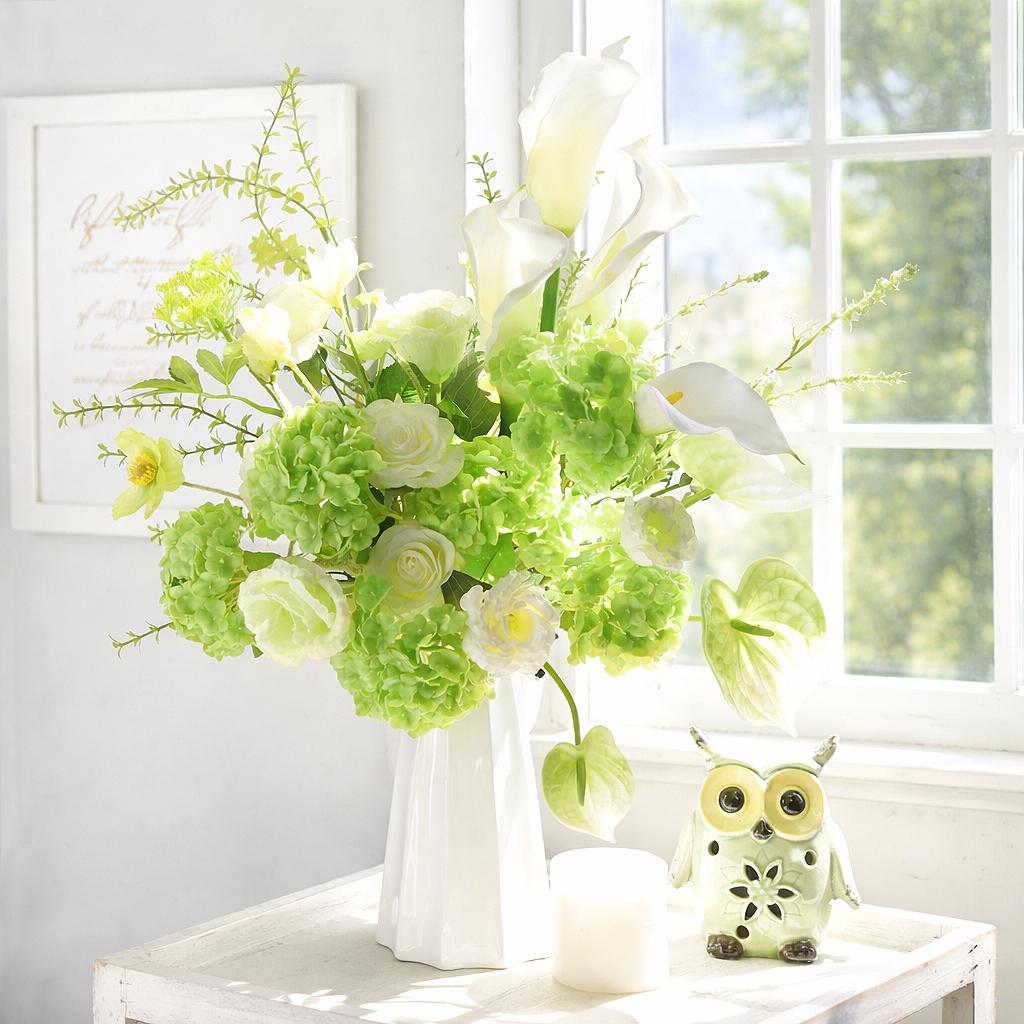 Lime green hydrangea stems arranged in a white vase on a bright tabletop near a window.