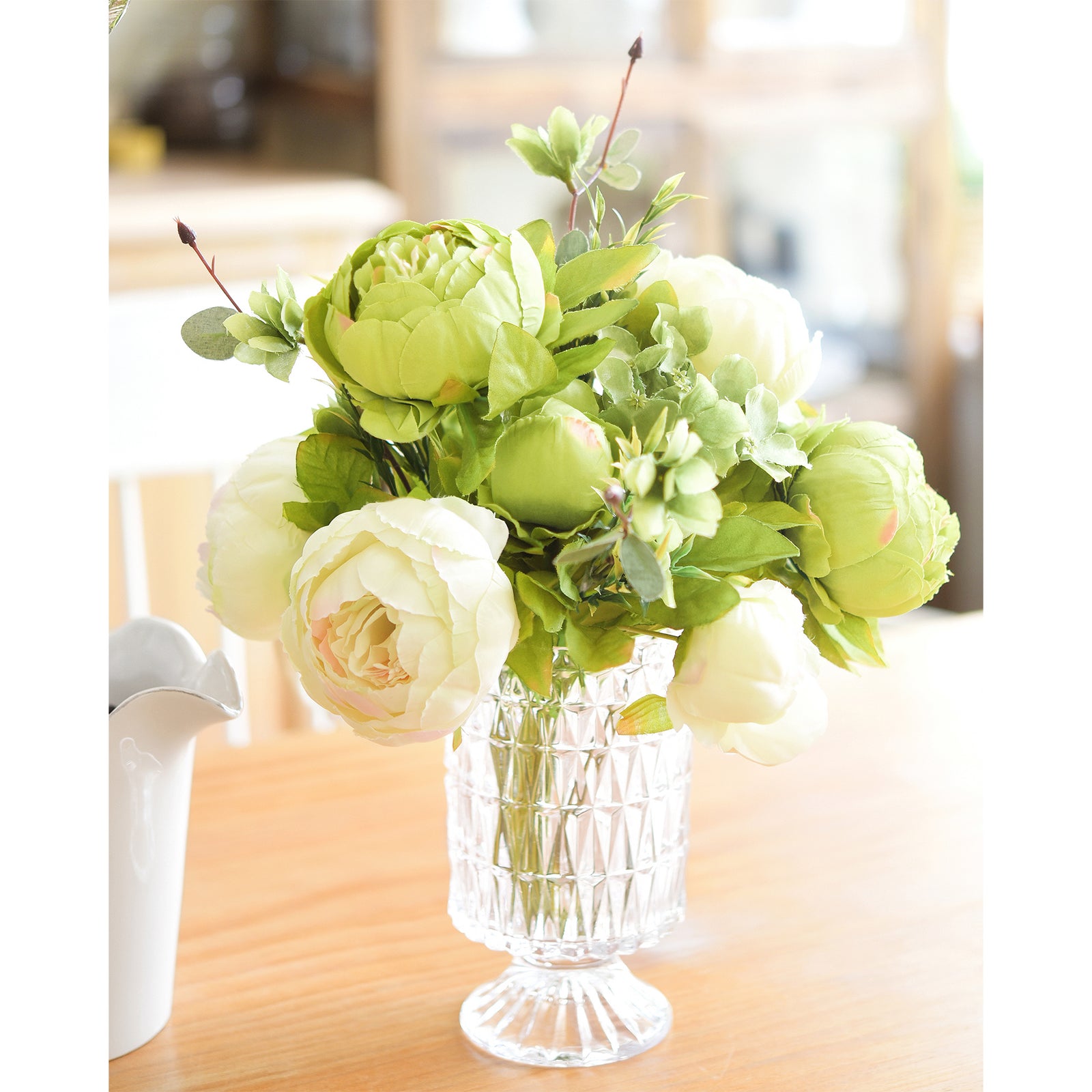 Moss green and cream faux peony bouquet arranged in a clear glass vase with candles for tabletop styling.