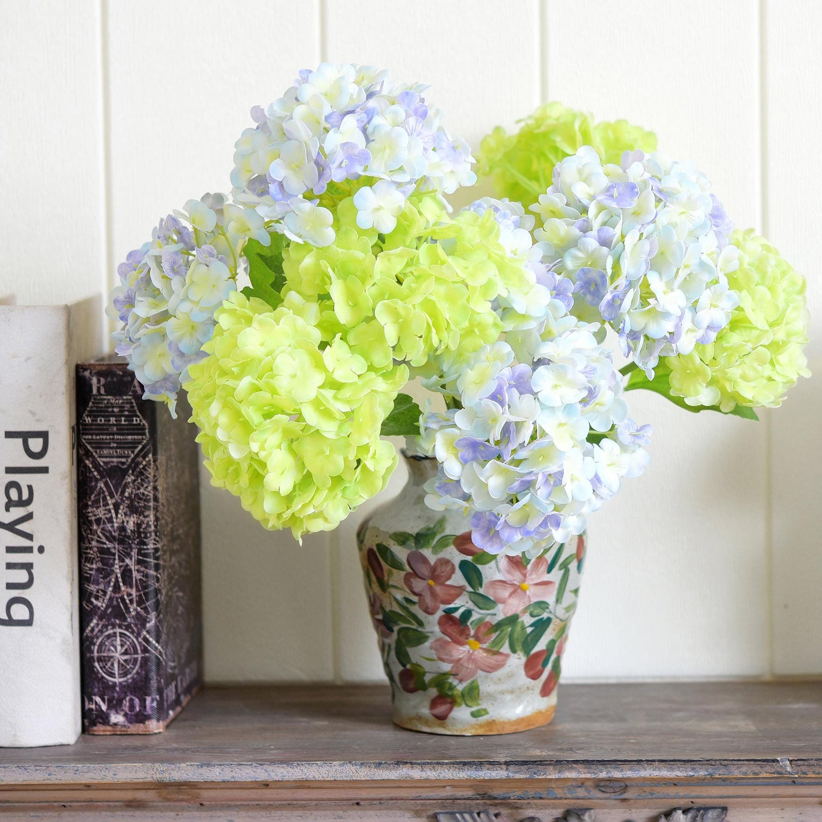 Periwinkle white and spring green hydrangea stems arranged in a patterned vase on a wooden shelf.