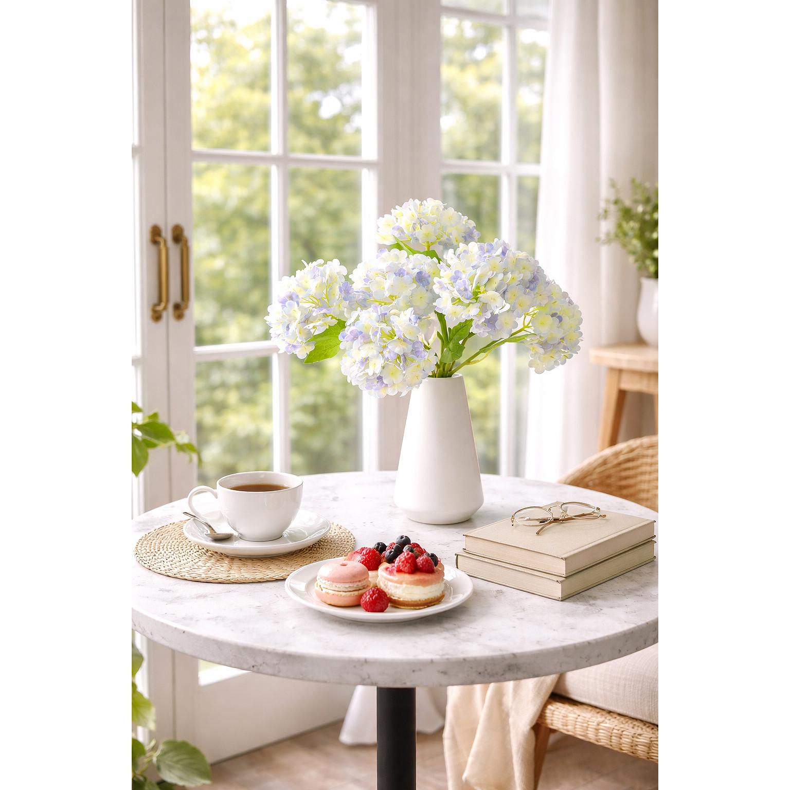 Periwinkle and white hydrangea stems arranged in a white vase by a bright window.