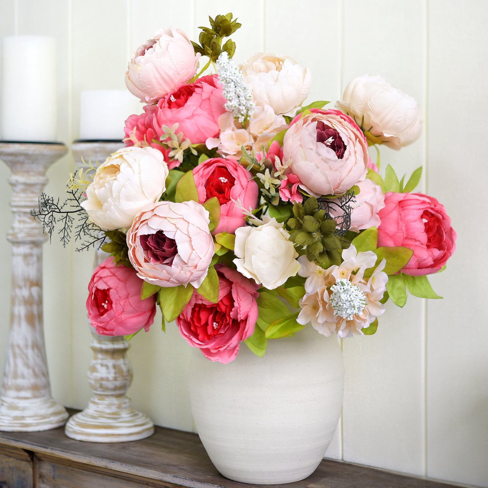 Artificial peonies in pink, blush, and ivory displayed in a white ceramic vase on a shelf with candles and frame for a romantic home setting.
