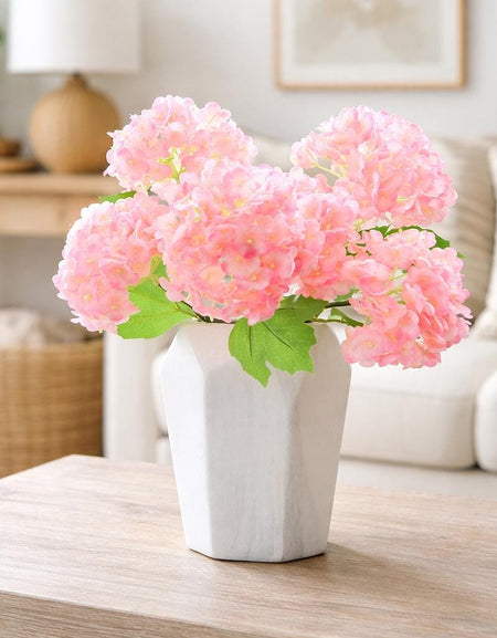 Pink real touch hydrangea stems arranged in a white vase on a light wood table.