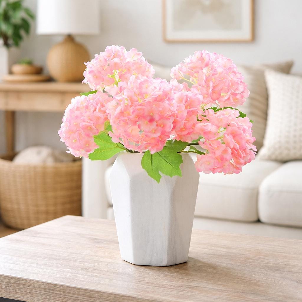 Pink real touch hydrangea stems arranged in a white vase on a light wood table.