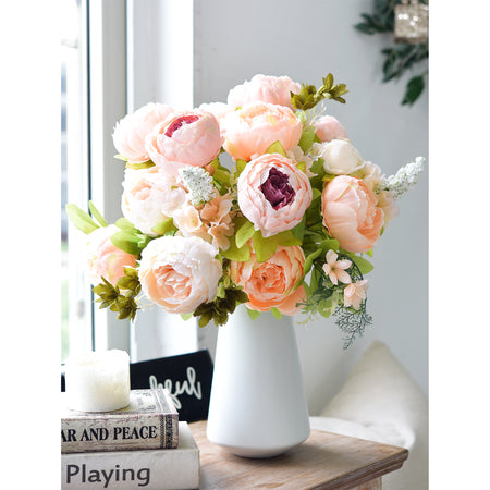 Elegant silk peony bouquet in a modern white vase displayed on a table by a window, featuring light peach and cream blooms with realistic greenery.