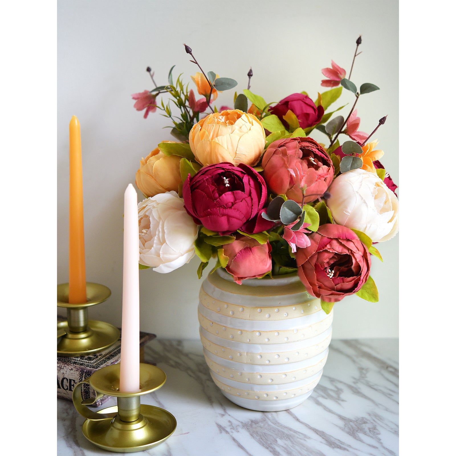 Silk peony and hydrangea bouquet in a textured ceramic vase, styled with two gold taper candle holders and pastel-colored candles on a marble surface.


