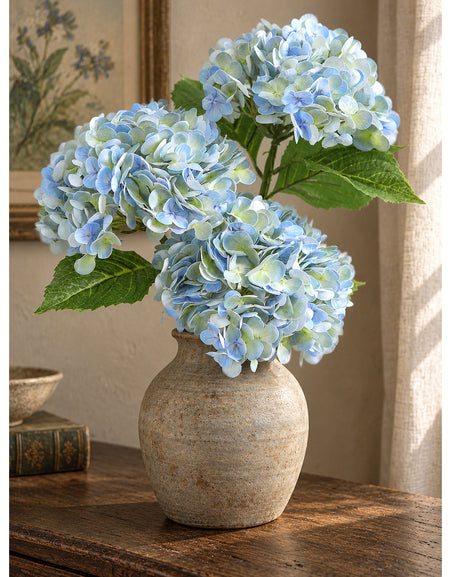 Three vintage blue green hydrangeas in a rounded stoneware vase beside books.