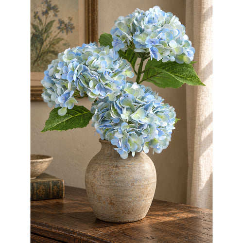 Three vintage blue green hydrangeas in a rounded stoneware vase beside books.
