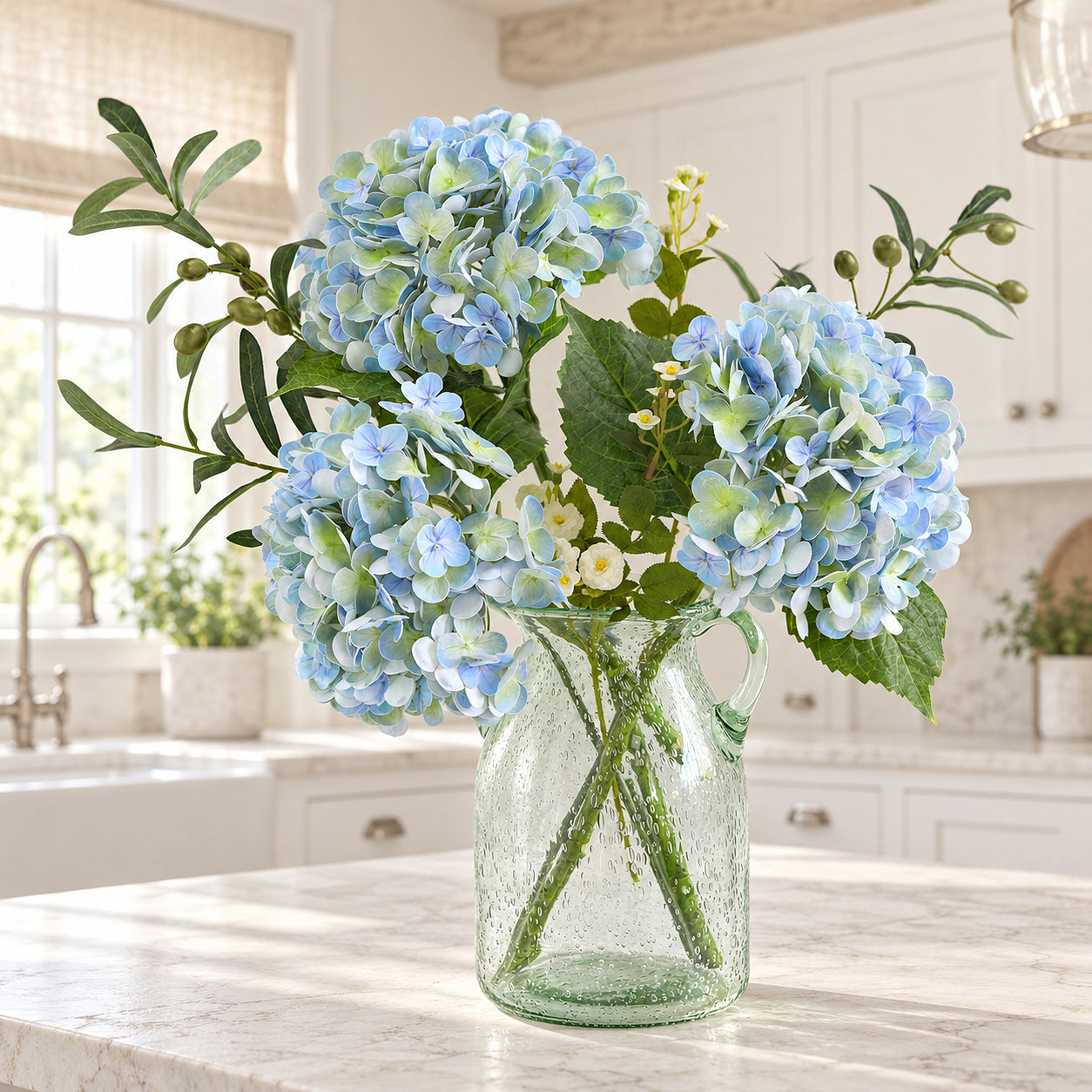 Three vintage blue green hydrangeas with greenery and small white flowers in a glass vase on a kitchen island.