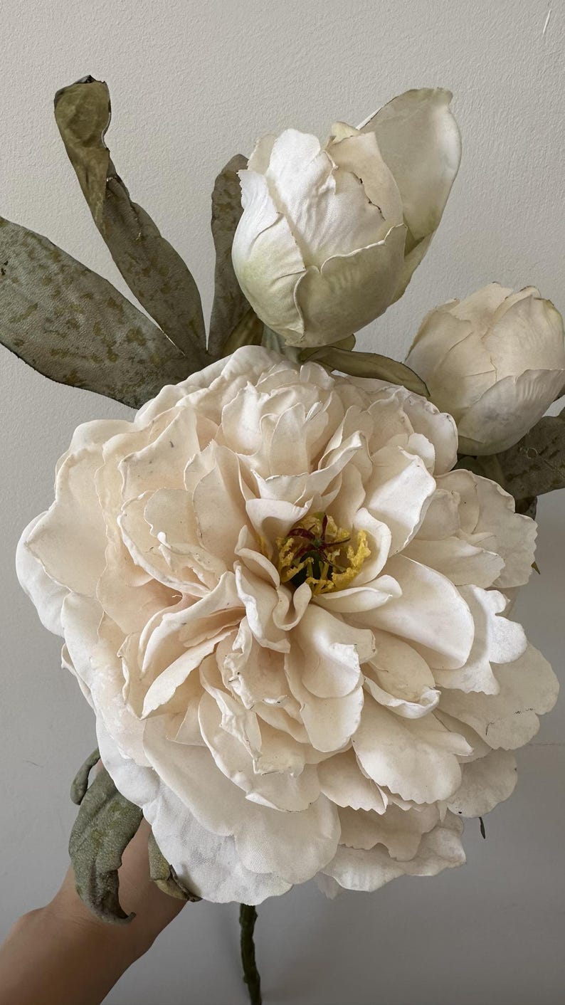 Close-up of an ivory artificial peony flower with a warm yellow center, two buds and mottled foliage.