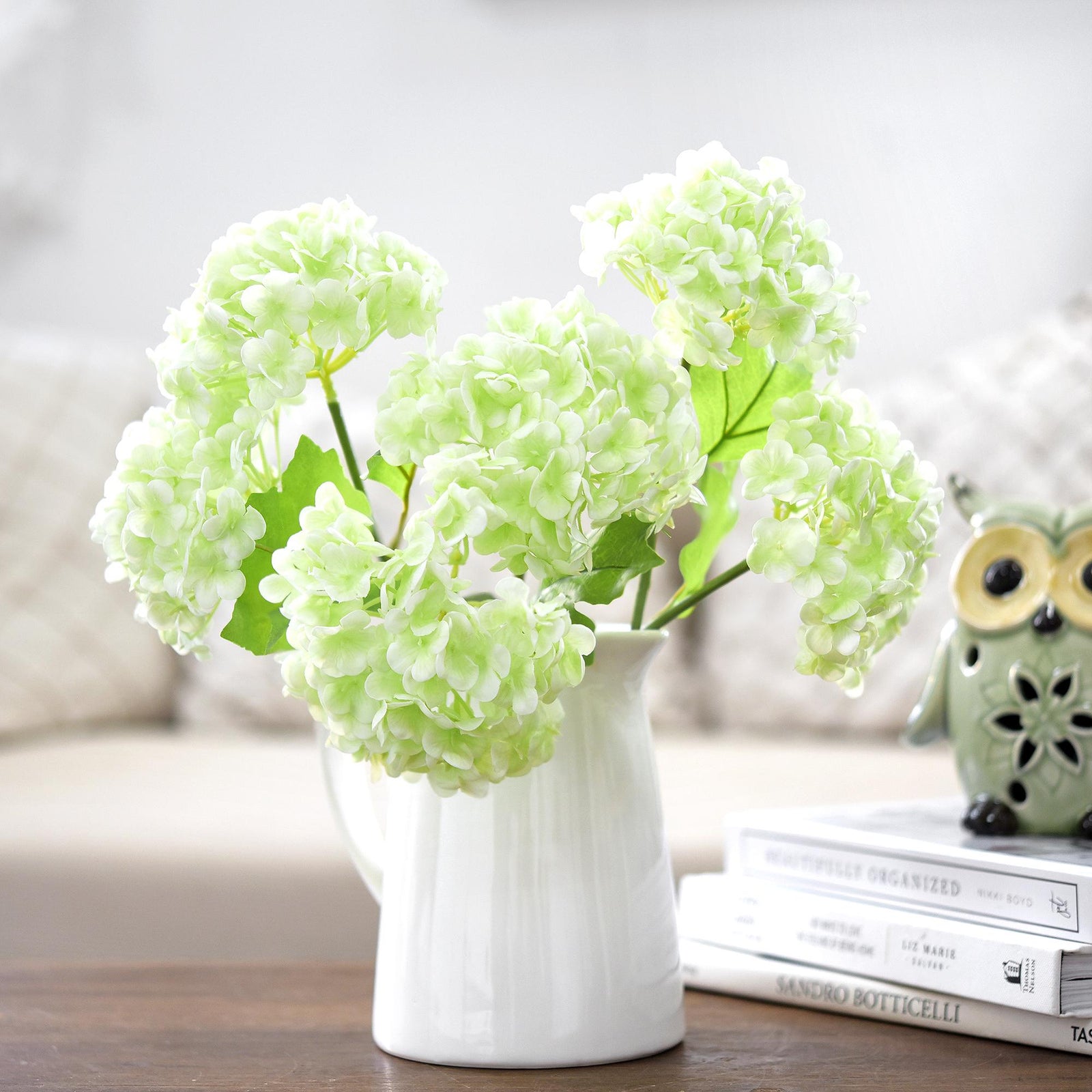 White and green real touch hydrangea stems arranged in a white vase on stacked books.