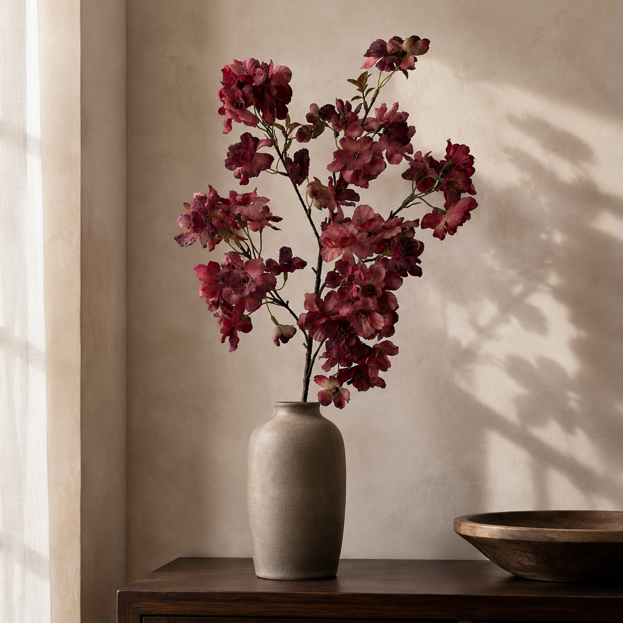 Wine burgundy artificial cherry blossom branch styled in a taupe vase on a dark wood console